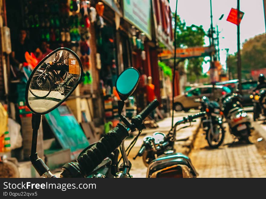 Several motorcycles parked on a street in India with a mirror on one of them showing what`s behind