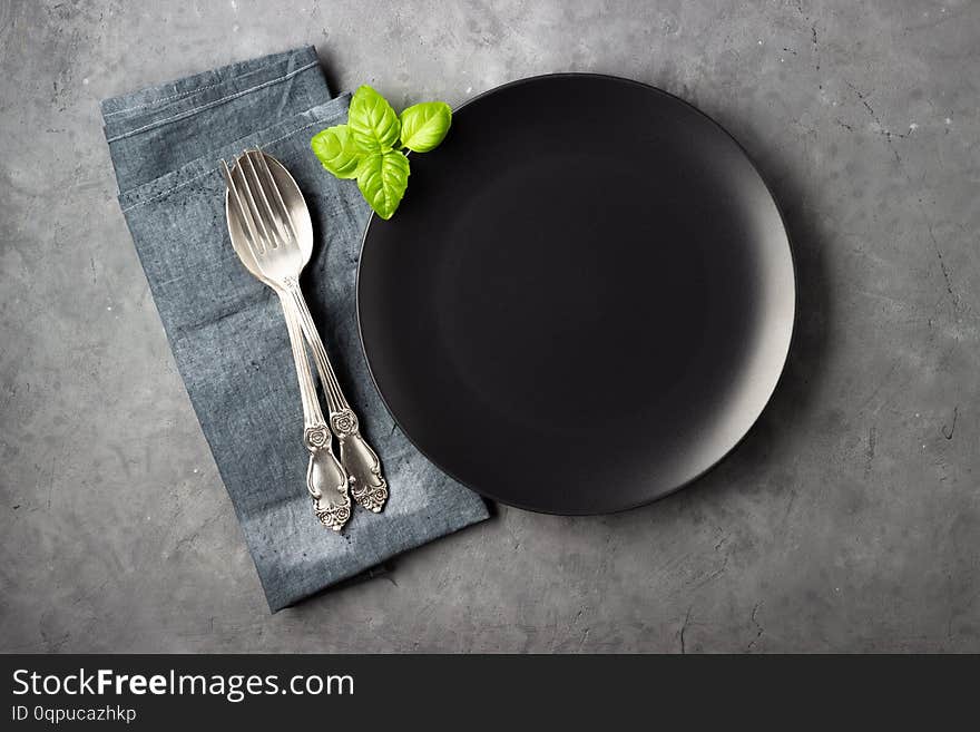 Table setting. Black plate, cutlery and napkin over gray concrete background