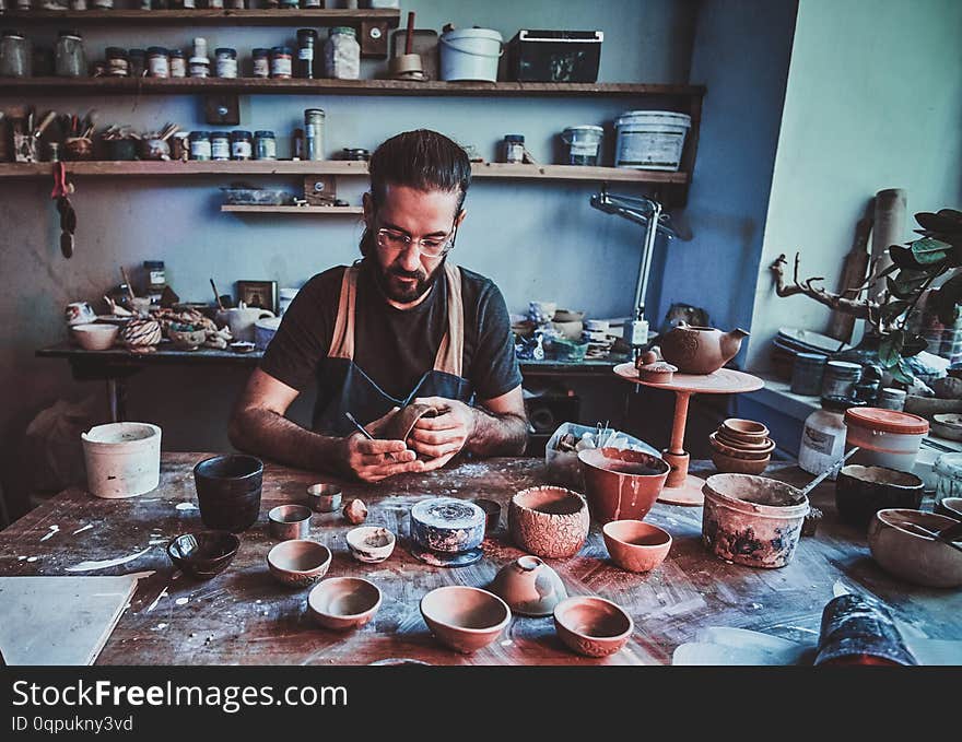 Pensive entusiastici potter in glasses at his own workshop is workig on a new handmade teapot.