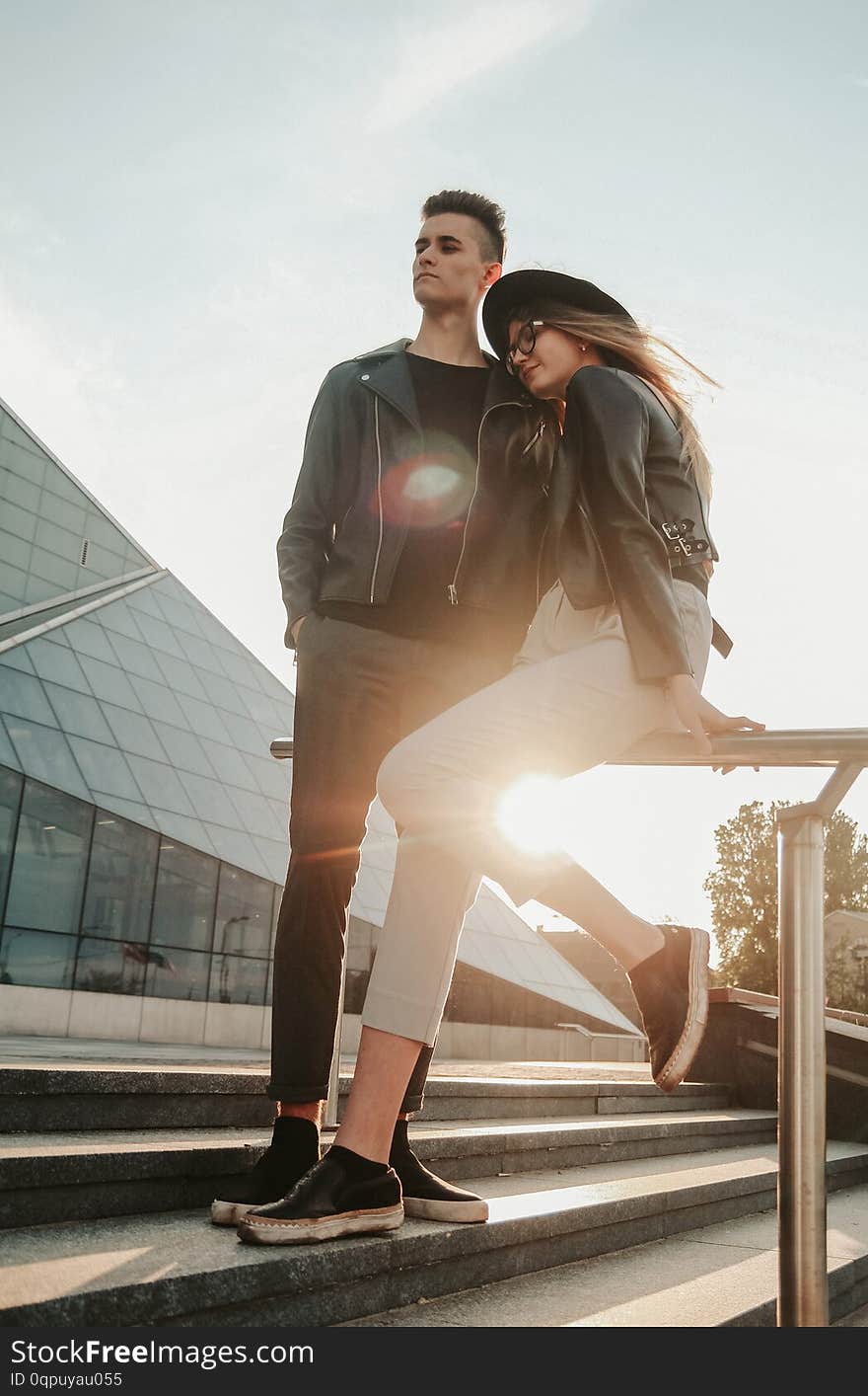 Young romantic couple is standing on stairs near glass building on sunrise time. They have casual style. Young romantic couple is standing on stairs near glass building on sunrise time. They have casual style.