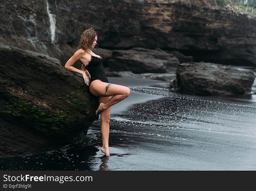 Sexy girl in swimsuit and tattoo on her leg poses on black sandy beach