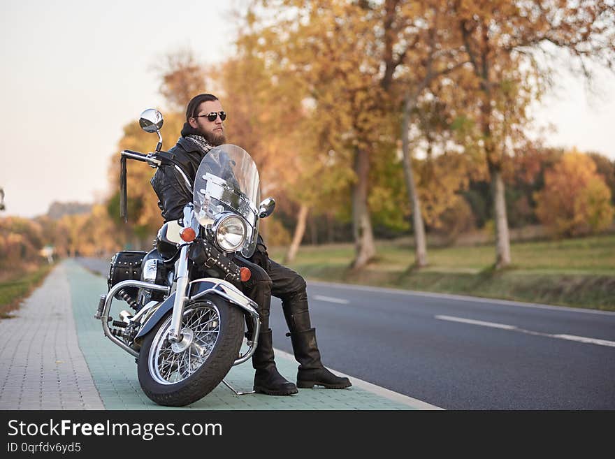 Young bearded biker in black leather clothing and dark sunglasses sitting on motorcycle on clean paved roadside, on background of empty straight asphalt road and vintage trees golden bokeh foliage. Young bearded biker in black leather clothing and dark sunglasses sitting on motorcycle on clean paved roadside, on background of empty straight asphalt road and vintage trees golden bokeh foliage.