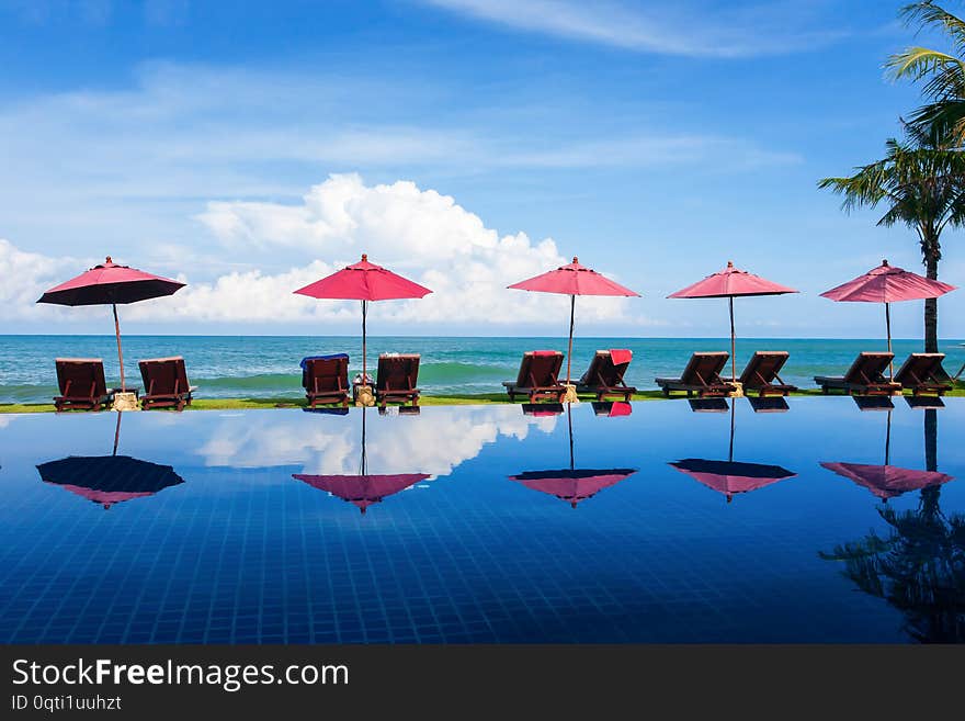 Scenery tropical resort with swimming pool near palm beach on sunny day, sunbathing chair and red umbrella near the sea. The beach is open to the public.