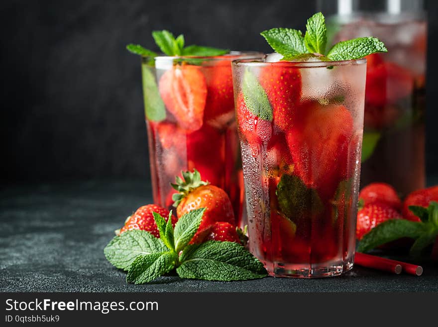 Fresh lemonade with ice, mint and strawberry on top in glass on black table background, copy space. Cold summer drink. Sparkling glasses with berry cocktail.