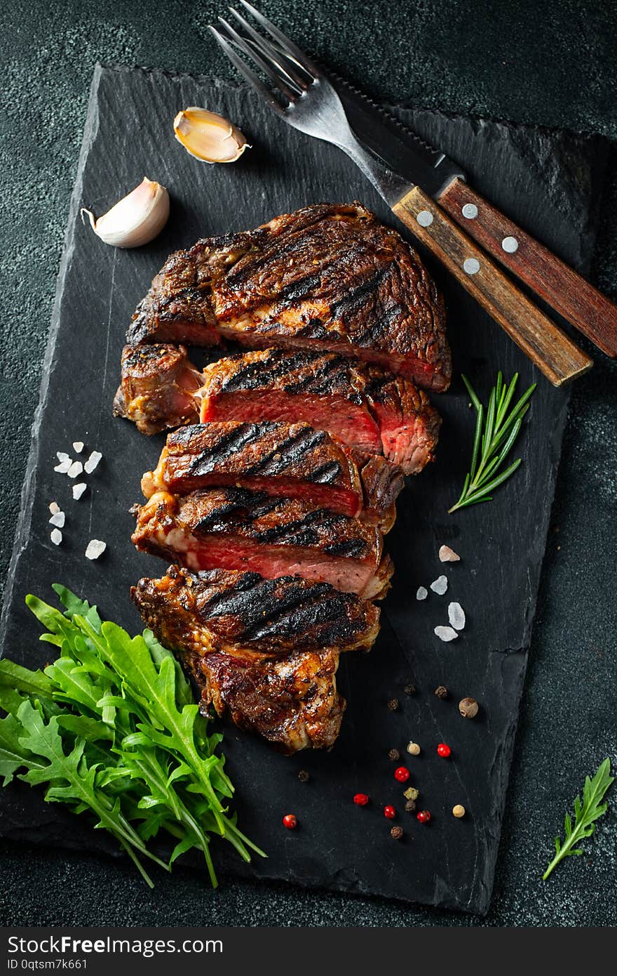 Sliced steak ribeye, grilled with pepper, garlic, salt and thyme served on a slate cutting Board on a dark stone background. Top view. Flat lay.