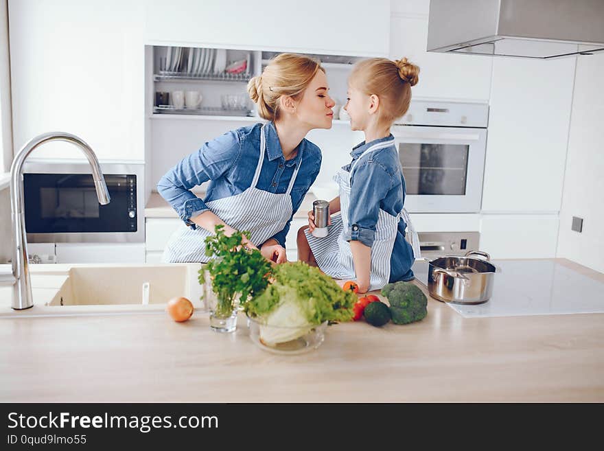 A young and beautiful mother in a blue shirt and apron is preparing a fresh vegetable salad at home in the kitchen, along with her little cute daughters with light hair. A young and beautiful mother in a blue shirt and apron is preparing a fresh vegetable salad at home in the kitchen, along with her little cute daughters with light hair