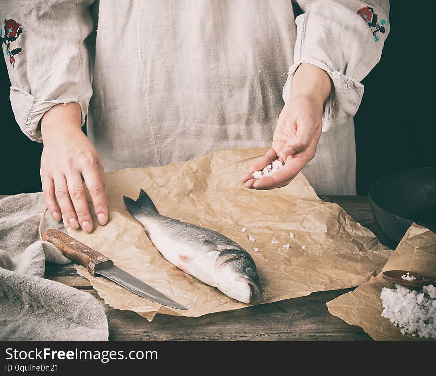 fresh whole sea bass fish lying on brown paper and a chef salt a product, black background