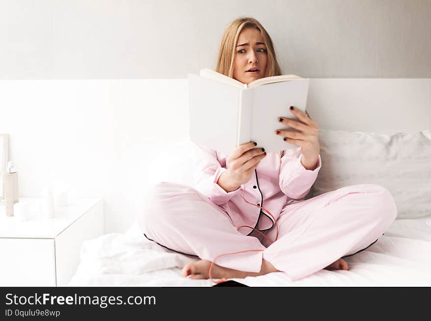 Young Woman Sitting On Bed And Amazedly Looking In Book At Home