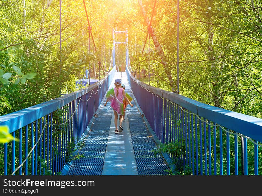 Two little happy girls walk along a hanging bridge on a sunny day.