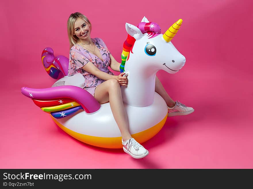 A beautiful blonde girl in a sexy sundress with slim legs in white sneakers sits on an inflatable multi-colored unicorn on a pink