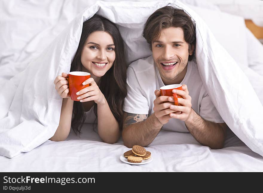 Good morning coffee. Cheerful couple hiding under duvet with cups and cookies, free space