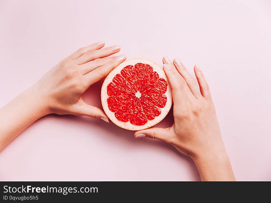 Female`s hands holding grapefruit on pink background with tropic plants and leaves. Top view, flat lay. Vegetarian food concept