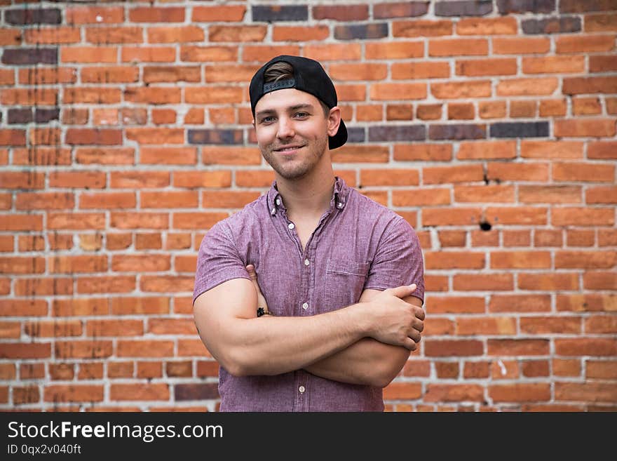 Handsome Young Caucasian Man with Cellphone and Backwards Hat Smiling for Portraits in Front of Textured Brick Wall Outside