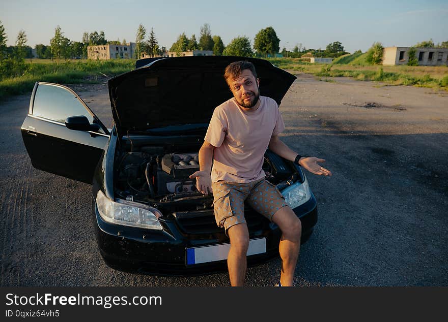 The guy in the pink t-shirt checks the engine of a black car lifting the hood in the country outside the road. The guy in the pink t-shirt checks the engine of a black car lifting the hood in the country outside the road