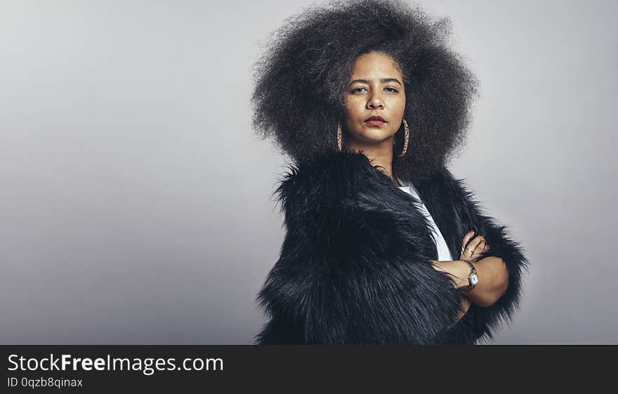 Portrait of woman in fur coat standing with her arms crossed looking at camera. Woman in afro hairstyle standing against grey background