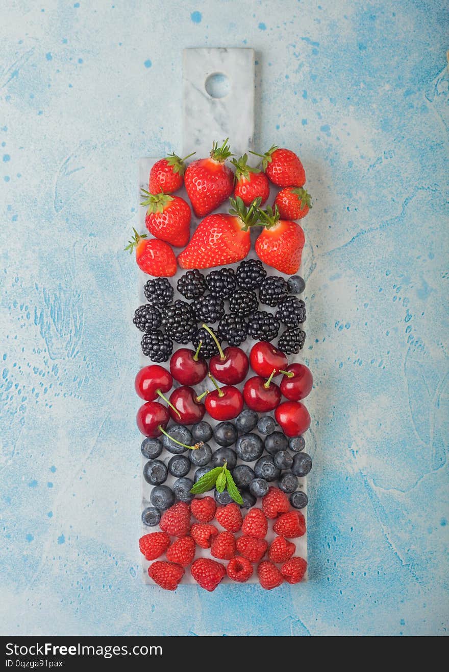 Fresh organic summer berries mix on white marble board on blue kitchen table background. Raspberries, strawberries, blueberries