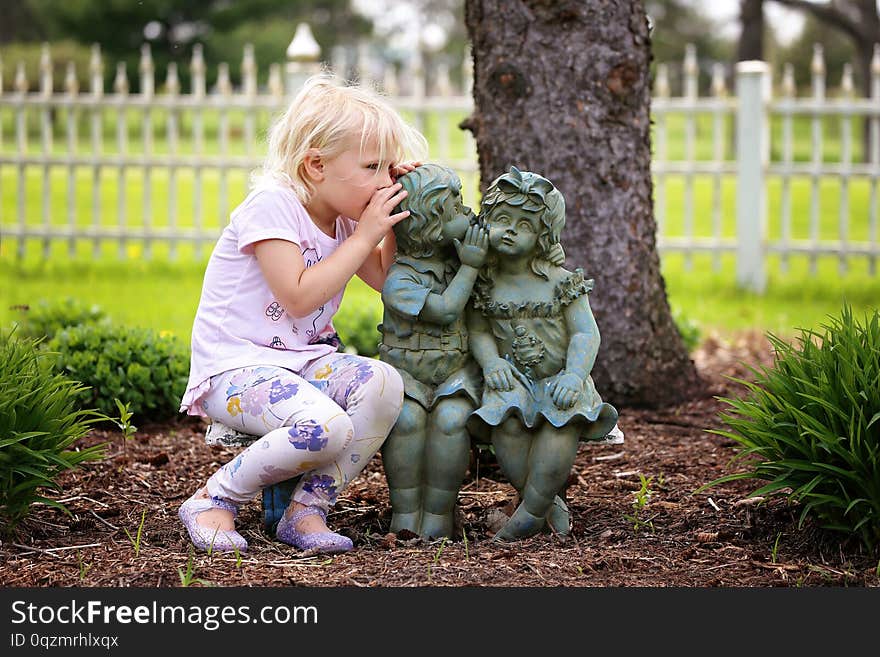 A cute little girl is sitting on a bench, whispering a secret as she plays with two little garden statue friends. A cute little girl is sitting on a bench, whispering a secret as she plays with two little garden statue friends