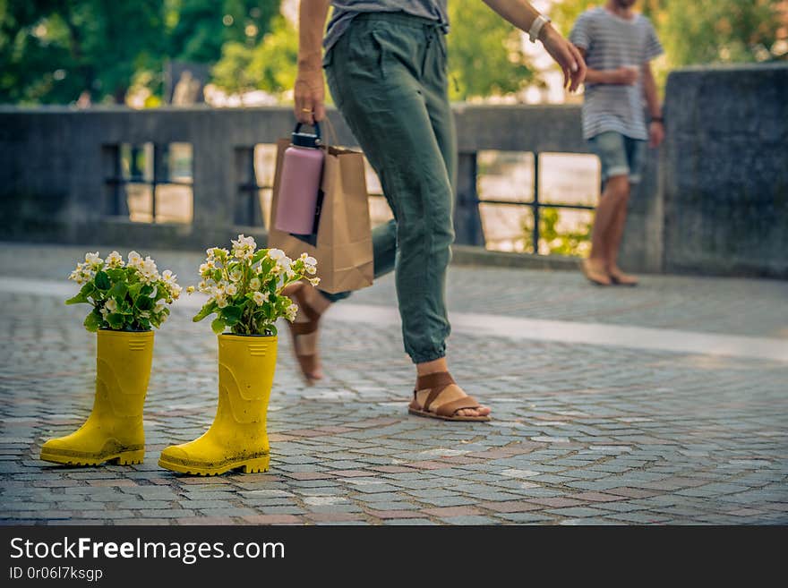 A pair of yellow rubber boots in the middle of a street in which flowers grow. in the background, blurry people walk by