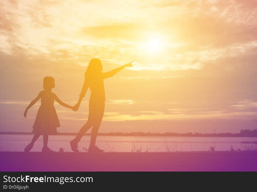 A silhouette of a happy young girl child the arms of his loving mother for a hug, in front of the sunset in the sky on a summer