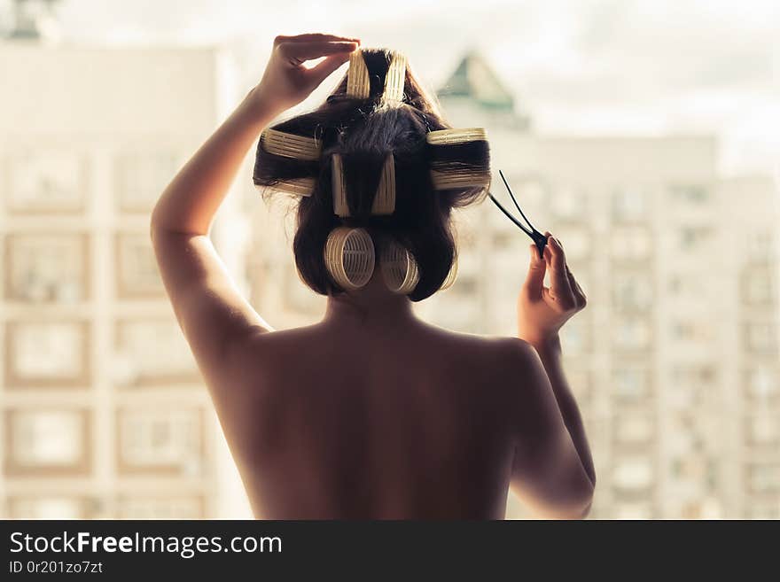 Naked young woman in a huge hair curlers standing near window opposite high building.