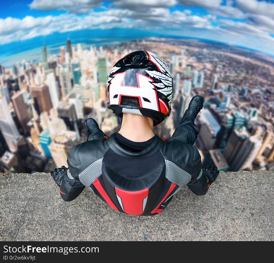 Motorcyclist in full gear and helmet sitting on a tall building. Man looks down on the city