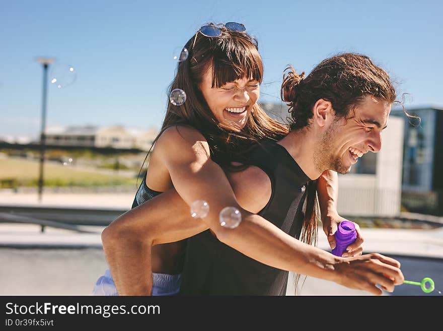 Couple having fun outdoors. Man piggybacking women playing with soap bubble outdoors