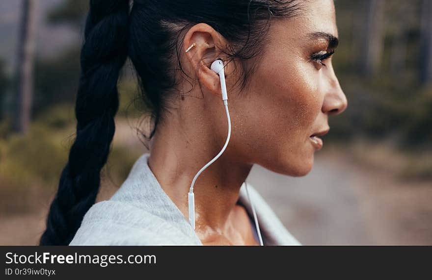 Side view of confident young woman with earphones standing outdoors after a morning run. Female athlete taking a rest after outdoor workout. Side view of confident young woman with earphones standing outdoors after a morning run. Female athlete taking a rest after outdoor workout