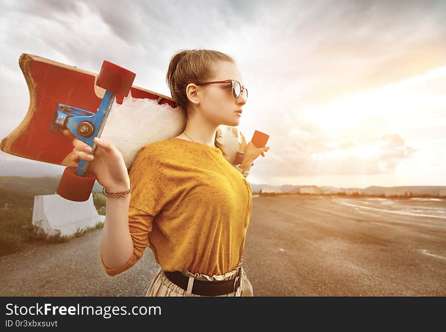 Beautiful and fashionable young woman in sunglasses and with a tattoo poses with a skateboard or longboard against the sunset sky.