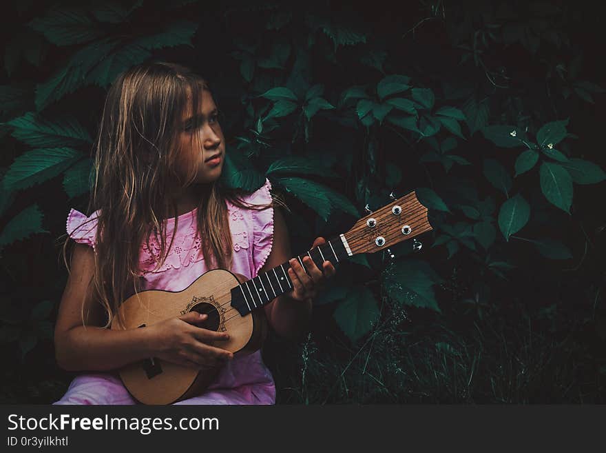 Beautiful young girl with blond hair wearing pink dress playing ukulele and dreaming on dark nature background. Grape leaves. Cutie lady learns play guitar concept. Beautiful young girl with blond hair wearing pink dress playing ukulele and dreaming on dark nature background. Grape leaves. Cutie lady learns play guitar concept