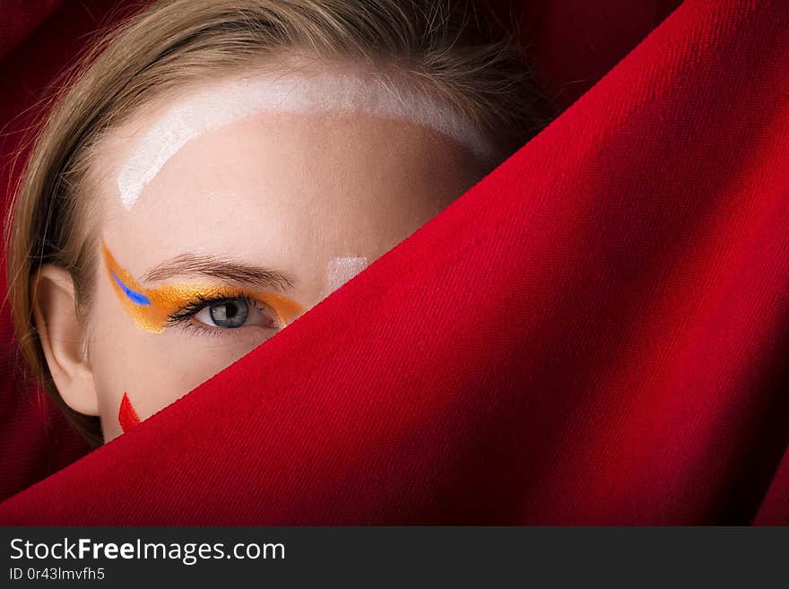 Girl with color make-up on a red background. A blonde with colored make-up peeks out because of the red cloth.