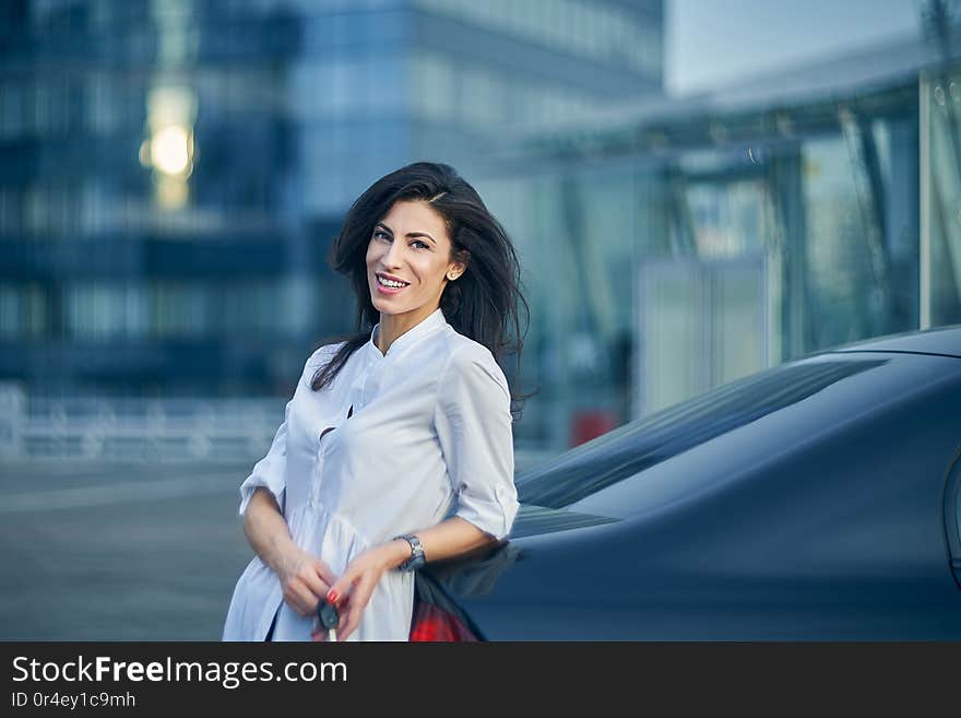 Smiling business woman standing outdoors leaning on the car with modern glass structure of office buildings at background. Smiling business woman standing outdoors leaning on the car with modern glass structure of office buildings at background