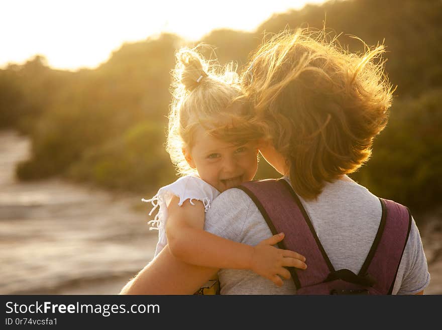 Portrait of a 3 year old Caucasian girl with blond hair who shows her tongue playfully. In the arms of the mother, backlit image at sunset