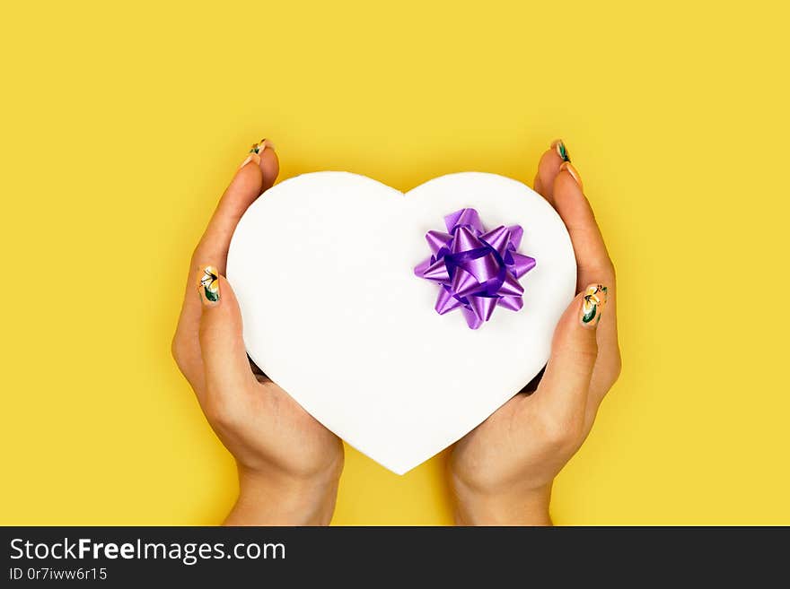 Heart shaped Valentines Day gift box in the hands of the girl on yellow paper background. Concept of the New Year, Christmas and Valentine`s Day.