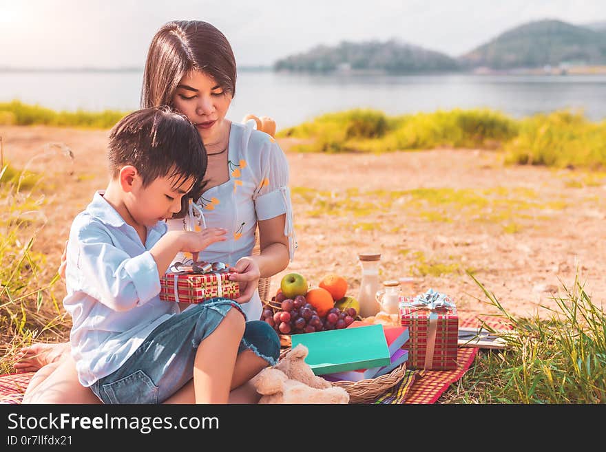 Beautiful Asian mother and son doing picnic and opening gift box from surprise in Birthday party on meadow near lake and mountain. Holiday and Vacation. People lifestyle and Happy family life concept