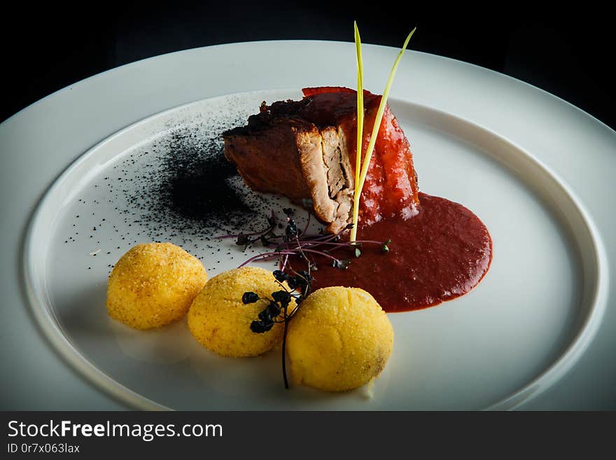 top view closeup roasted meat and potato balls decorated with spicy red sauce and dry herbs on white plate over black. top view closeup roasted meat and potato balls decorated with spicy red sauce and dry herbs on white plate over black