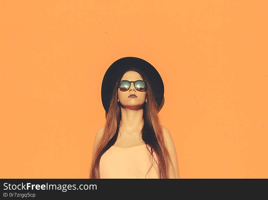 Fashionable girl standing wearing fashionable sunglasses and black hat with a solid color as background