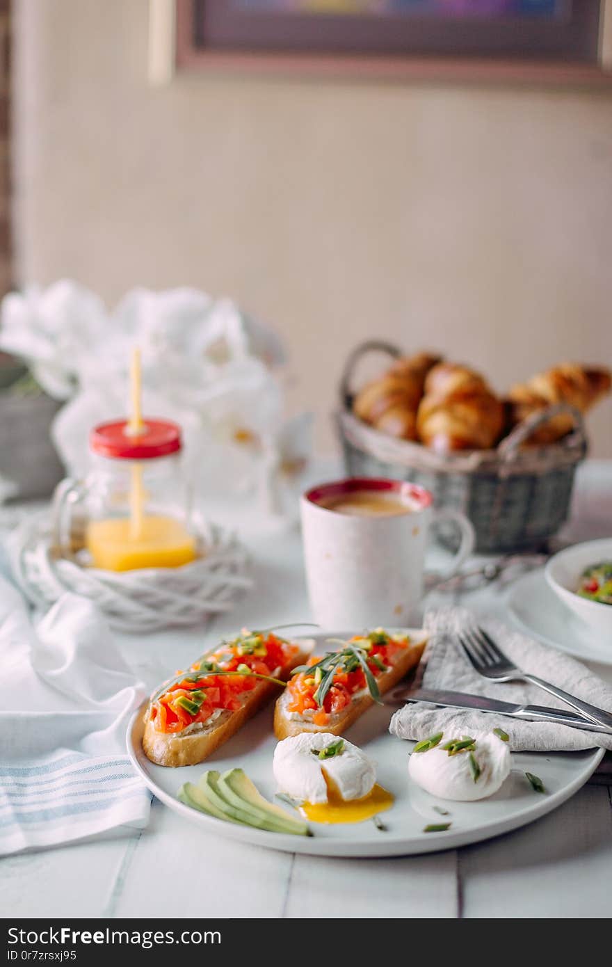 Photo of norway breakfast. Toasts with salmon and cream-cheese, boiled eggs served om a white plate standing on a white wooden table with vegetable salad, coffee, jar of orange juice and croissants on a background. Photo of norway breakfast. Toasts with salmon and cream-cheese, boiled eggs served om a white plate standing on a white wooden table with vegetable salad, coffee, jar of orange juice and croissants on a background.