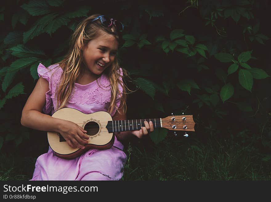 Beautiful young girl with blond hair wearing pink dress playing ukulele and smiling on dark nature background. Grape leaves. Cutie lady learns play guitar concept. Beautiful young girl with blond hair wearing pink dress playing ukulele and smiling on dark nature background. Grape leaves. Cutie lady learns play guitar concept