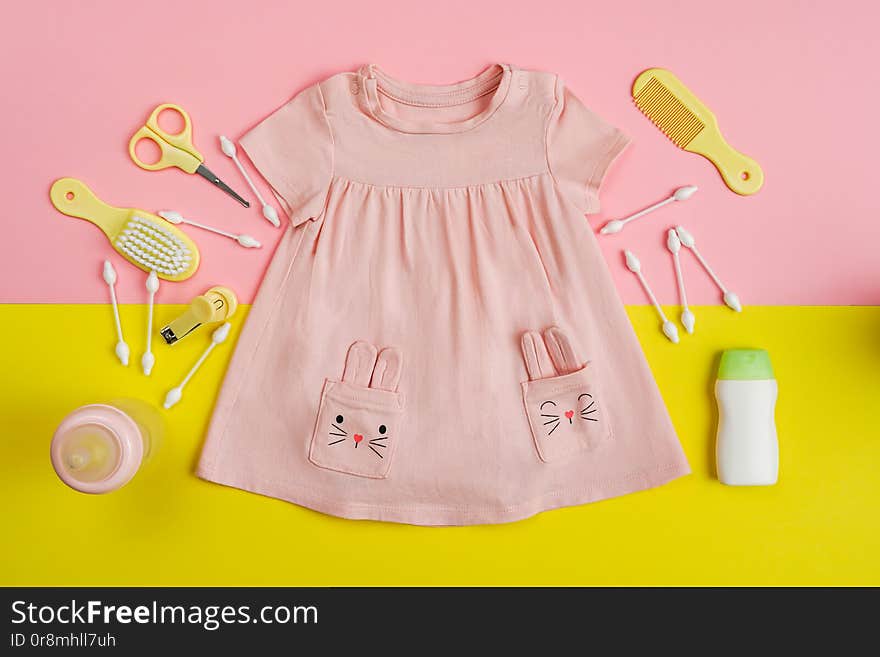 Baby girl dresseeding and necessities for baby. Milk bottle, shower gel, cotton buds, nail clippers and baby girl pink dress on yellon and pink background, top view.