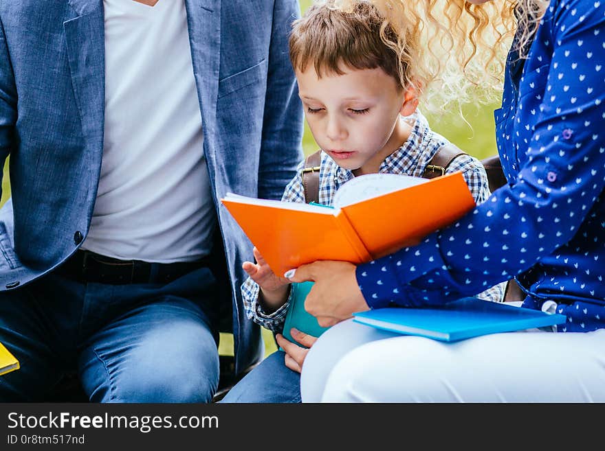 Engaged child boy sitting between mother and father and reading interesting book outdoor.