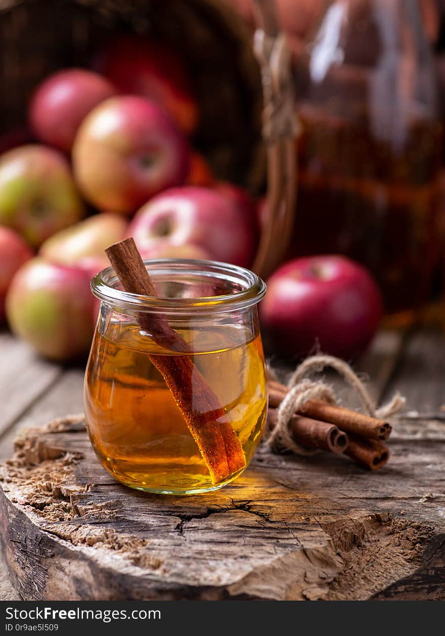 Glass of apple cider with cinnamon sticks on a rustic wood surface with red apples in background