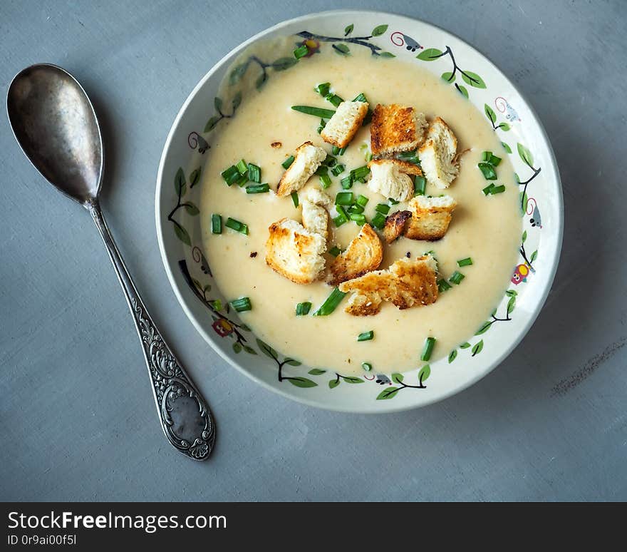 Light soup of mashed potatoes with cheese and fresh vegetables in a deep plate, shot from close range, near a tablespoon on a gray background