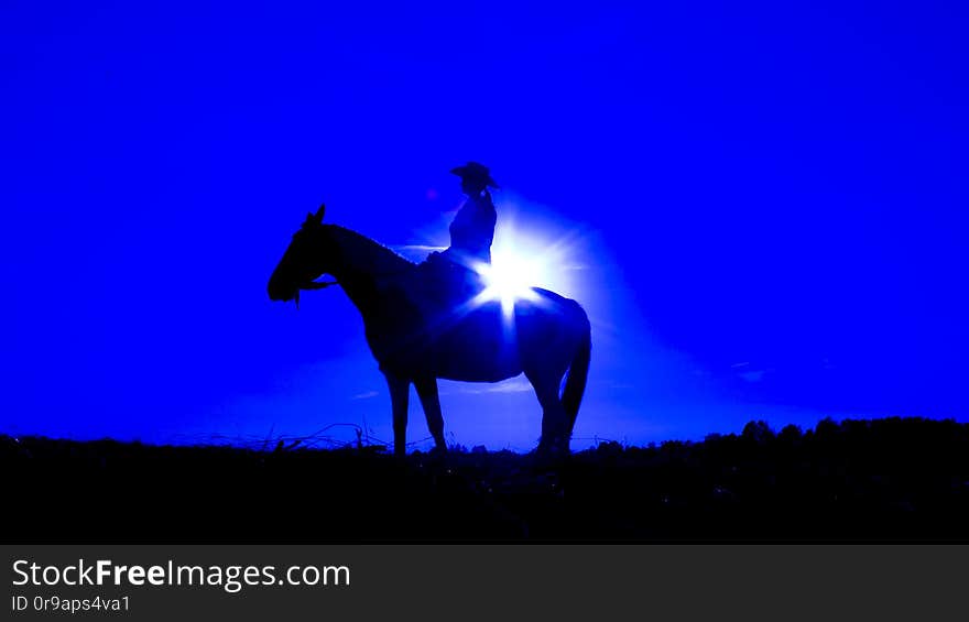 Silhouette of a cowgirl on open plain in western  riding a horse lit by the setting sun in blue. Silhouette of a cowgirl on open plain in western  riding a horse lit by the setting sun in blue