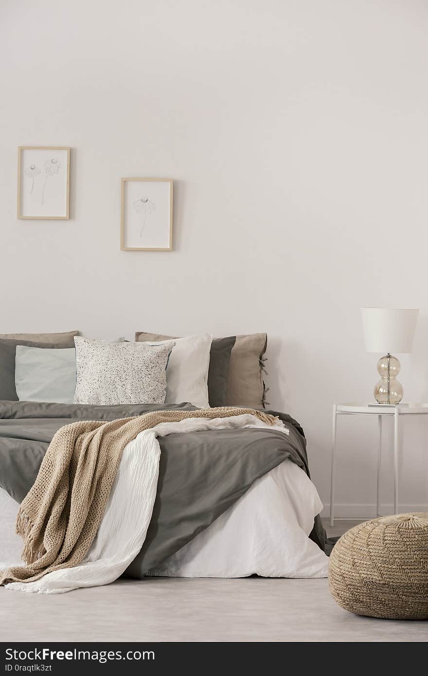 Stylish white lamp on simple nightstand table next to warm bed with cozy grey bedding