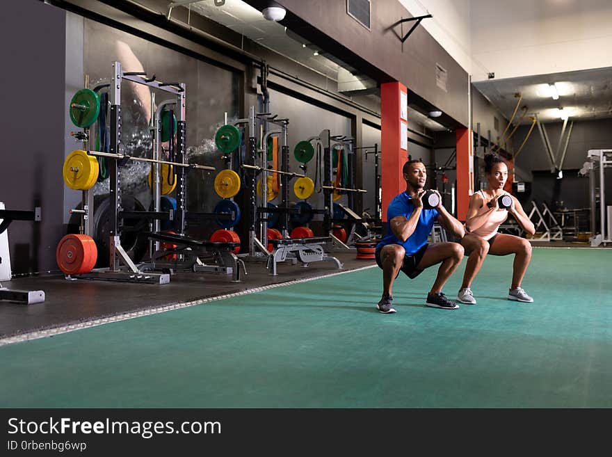 Side view of an African-American man and woman lifting ball weights with both arms while doing squats inside a room at a sports centre. Side view of an African-American man and woman lifting ball weights with both arms while doing squats inside a room at a sports centre