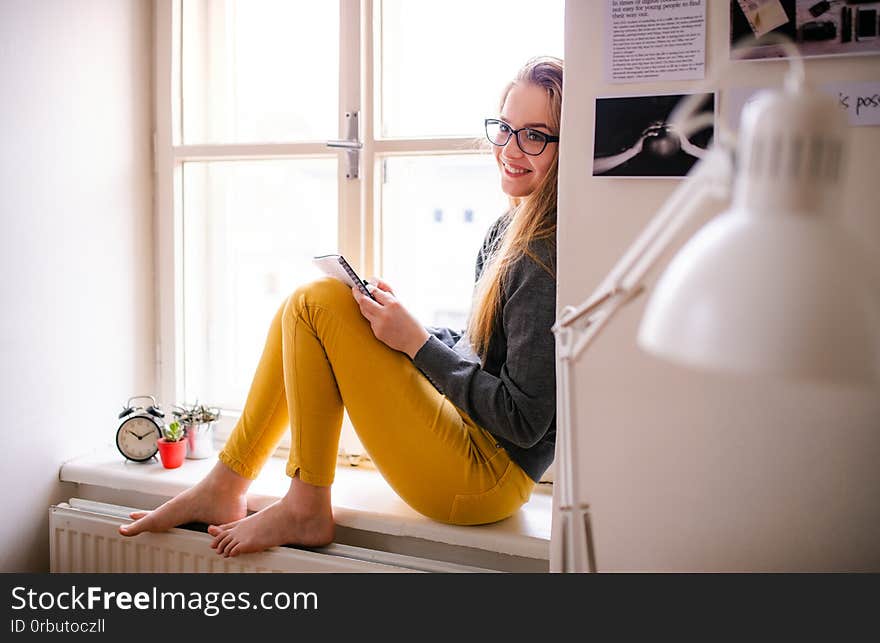 A young happy college female student with an exercise book sitting on window sill at home, studying. A young happy college female student with an exercise book sitting on window sill at home, studying.