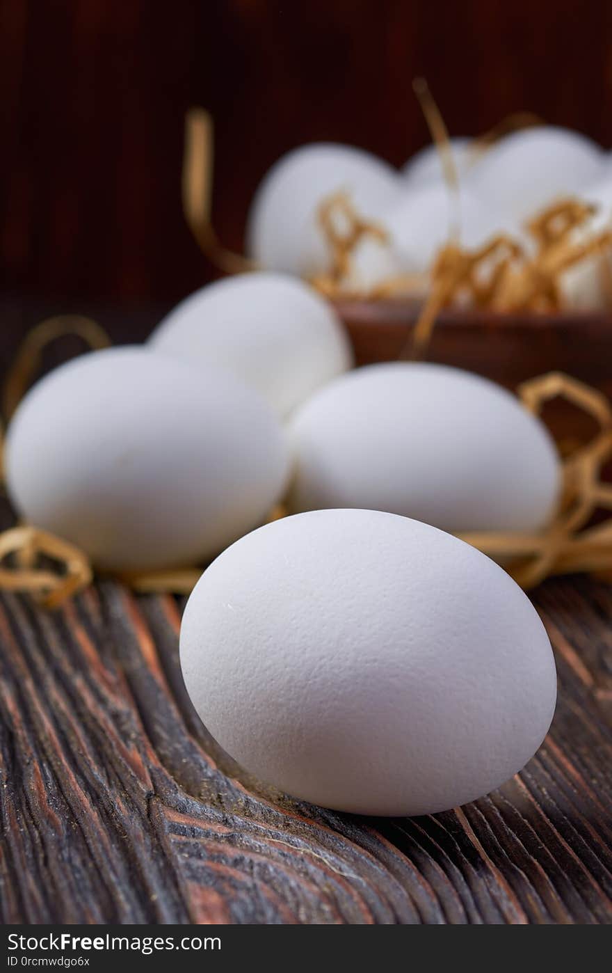 White eggs on a wooden table. In the background are eggs in a wooden bowl and on paper straw. Close up. Low key. White eggs on a wooden table. In the background are eggs in a wooden bowl and on paper straw. Close up. Low key