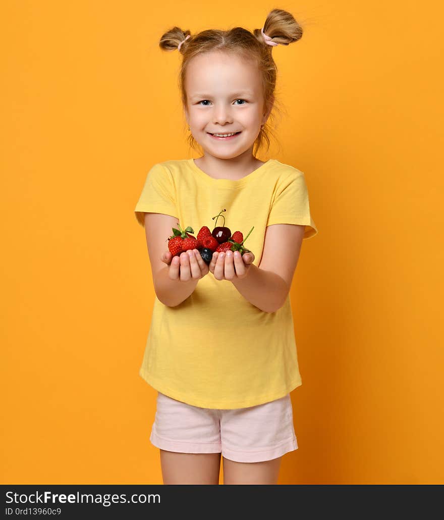 Baby girl kid blonde with funny buns demonstrates a handful of berries fruits in her palms hands and smiles satisfied with her harvest on yellow  background. Baby girl kid blonde with funny buns demonstrates a handful of berries fruits in her palms hands and smiles satisfied with her harvest on yellow  background