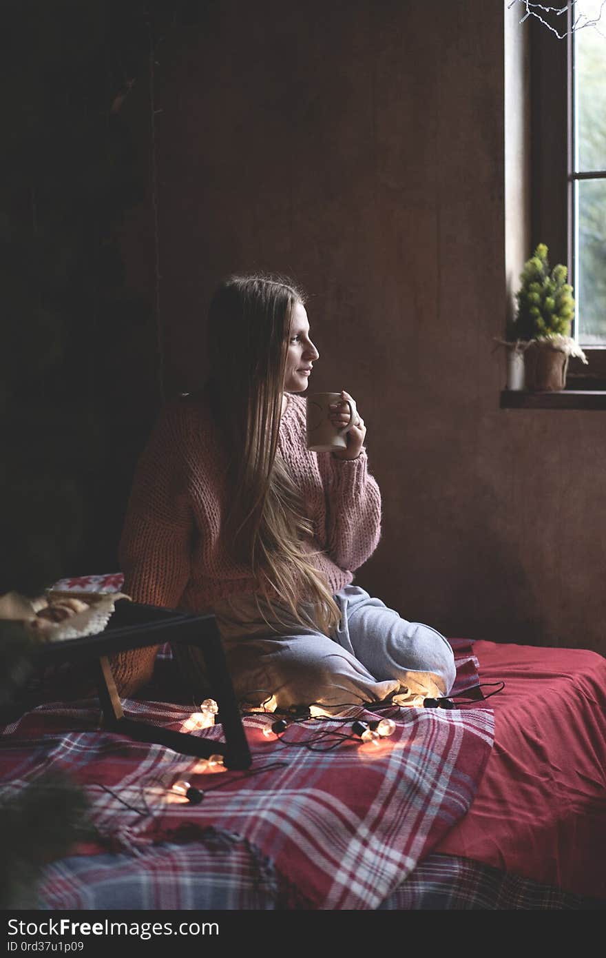 Caucasian young woman with long straight hair in a knitted sweater on the bed looks out the window and holds a mug with a drink. Christmas interior decor.