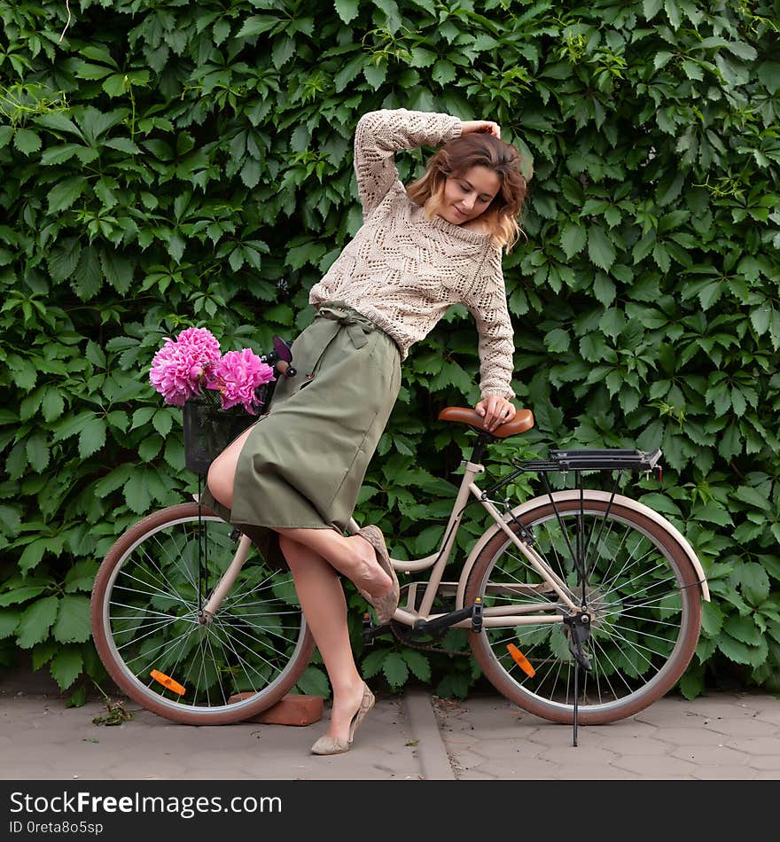 A young dark-haired woman in sweater and a beige skirt  sitting on beige retro bicycle  with bouquet of pink peonies in a wicker basket on the background of a vine-covered wall. A young dark-haired woman in sweater and a beige skirt  sitting on beige retro bicycle  with bouquet of pink peonies in a wicker basket on the background of a vine-covered wall